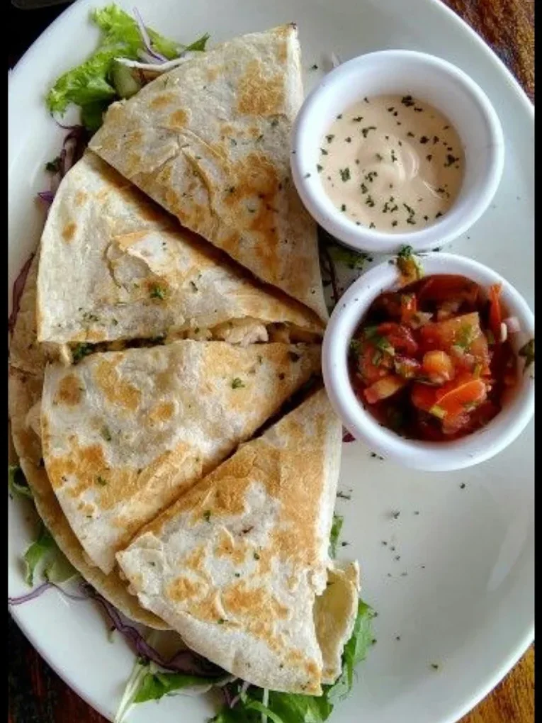 A flour quesadilla cut into wedges on an oval plate over mixed greens and red cabbage, with a cup of crema and a cup of pico de gallo.