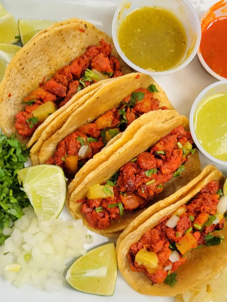 Four corn tortilla tacos on a white plate with red chile marinated meat and pineapple, beside chopped onion, cilantro, lime wedges and four cups of salsa.