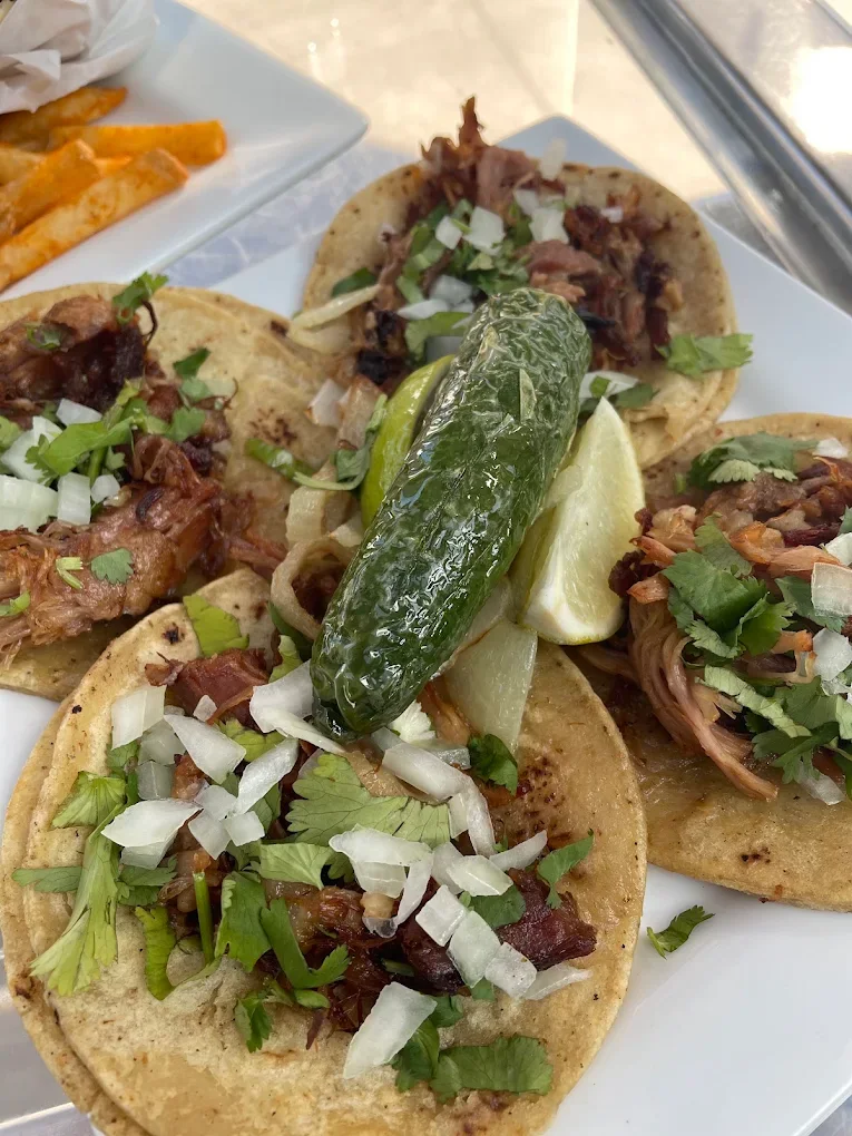 Four corn tortilla tacos of chopped pork topped with white onion and cilantro, a whole blistered jalapeno and lime wedges across the middle.