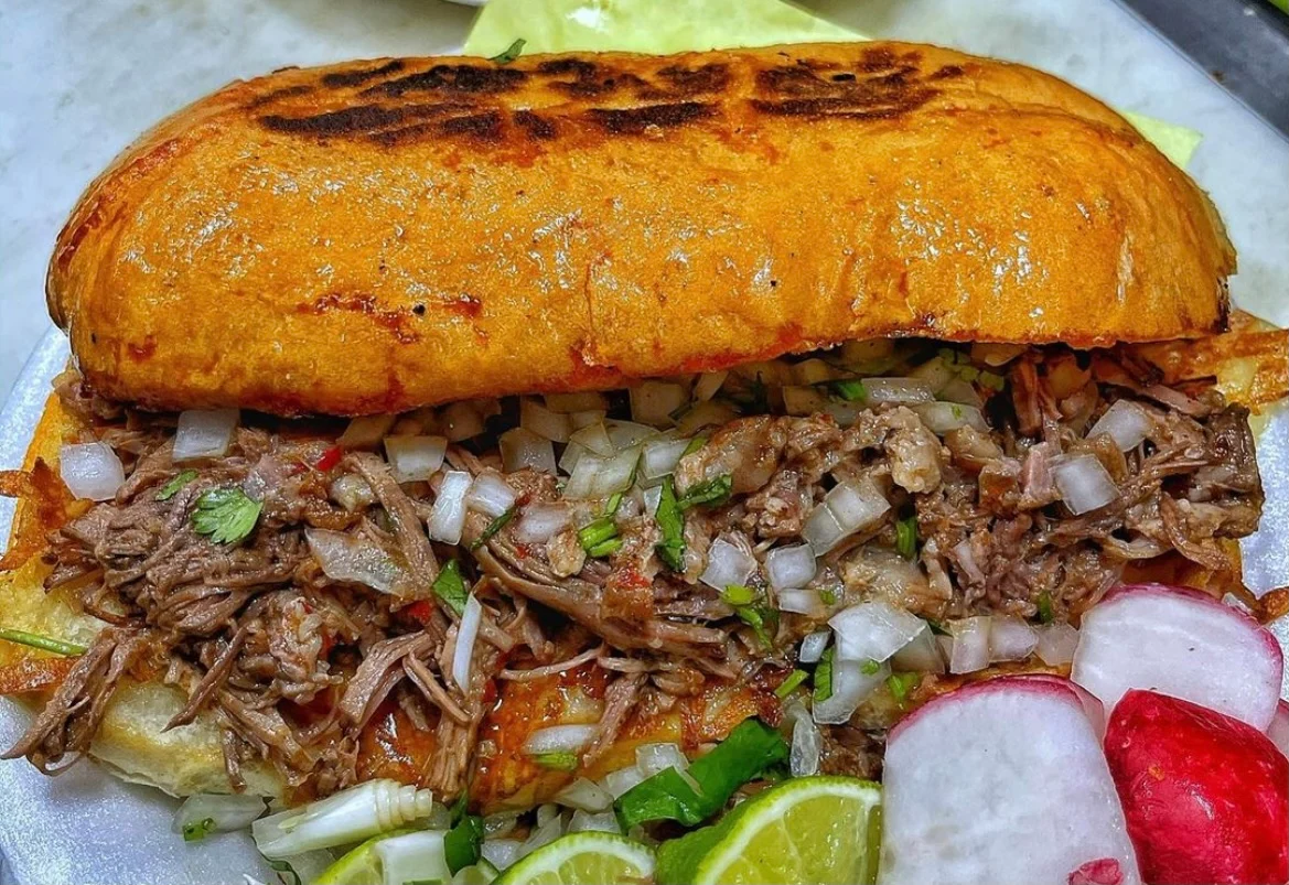 A torta on toasted telera bread piled with shredded meat, white onion and cilantro, with sliced radish and lime alongside.