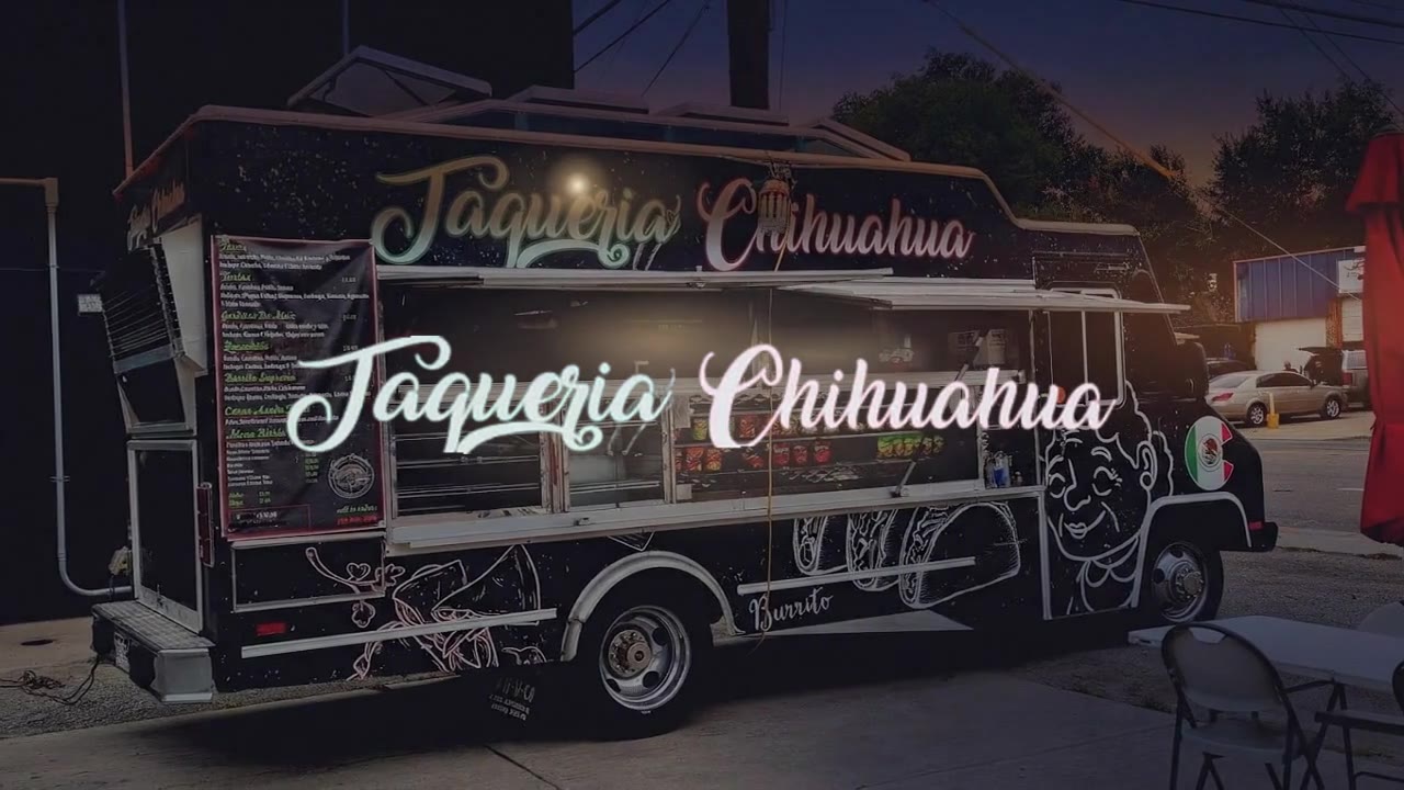 The Taqueria Chihuahua truck at dusk, matte black with its name painted in script, the service window glowing amber against an orange and indigo sky.