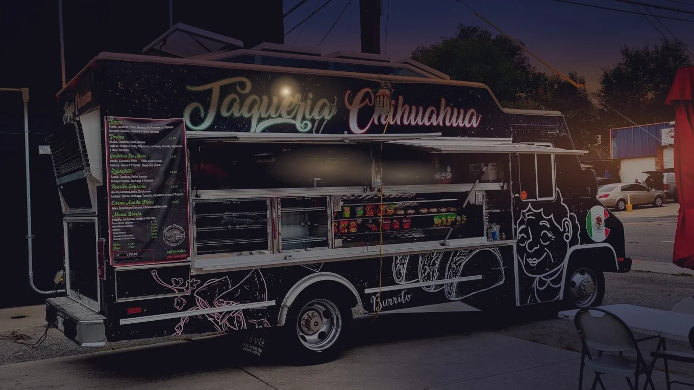 The Taqueria Chihuahua truck parked at dusk, the menu board and service window lit from inside, the painted name running along the side.
